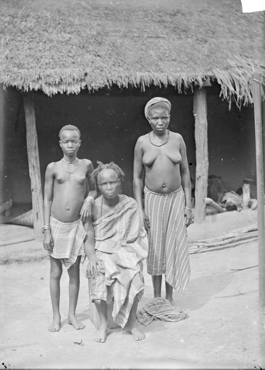 Three Igbo women and girls in striped woven fabrics in front of a thatched building.