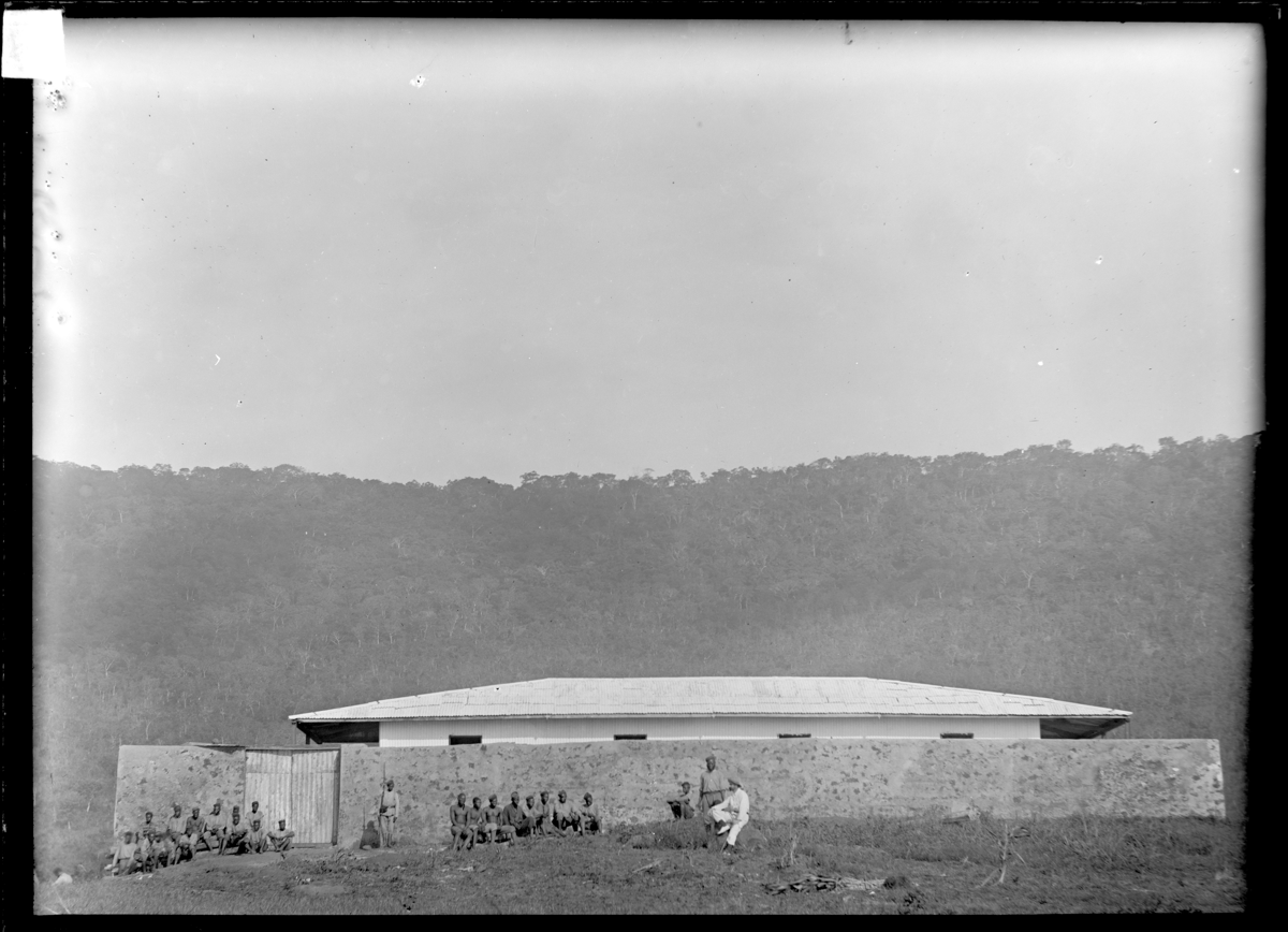 A photograph showing a group of people in simple clothing seated and standing before a masonry wall and building with a forested hill in the background.