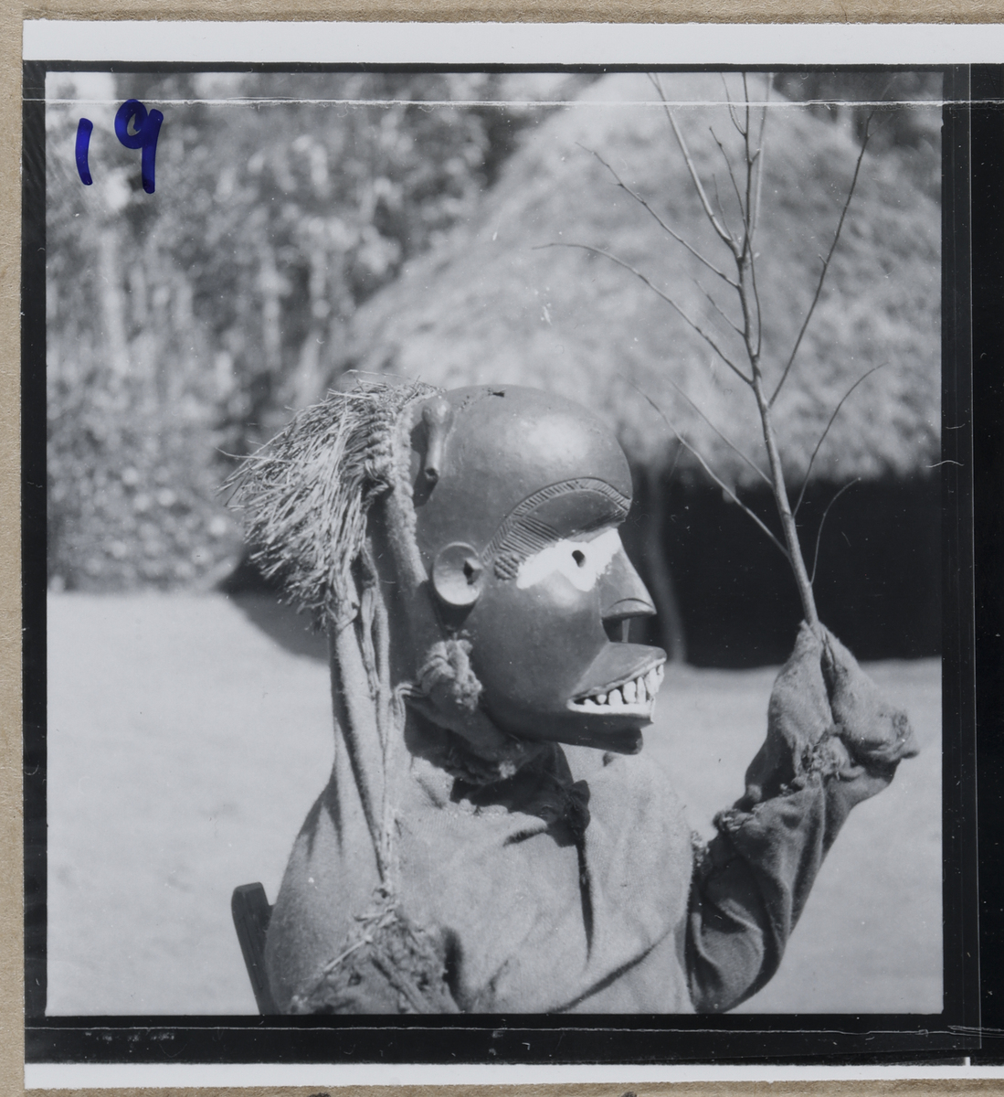 Profile view of an Iro Ekpetu masquerader wearing a black wooden mask with white painted accents and raffia hair.
