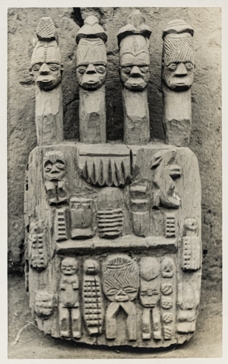 A photograph of a carved wooden board staff featuring relief figures and four projecting heads.