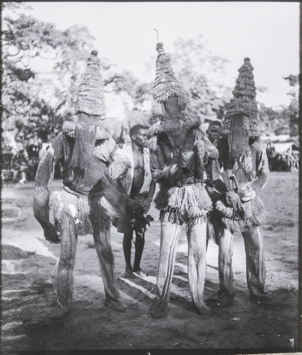 Three Onu Kamma masqueraders with large conical fiber masks and gongs, surrounded by onlookers