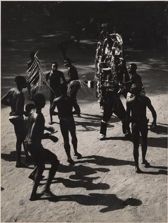 A photograph showing a group of Igbo men in a masquerade, one wearing an elaborate mask with streamers and rattles on his knees.