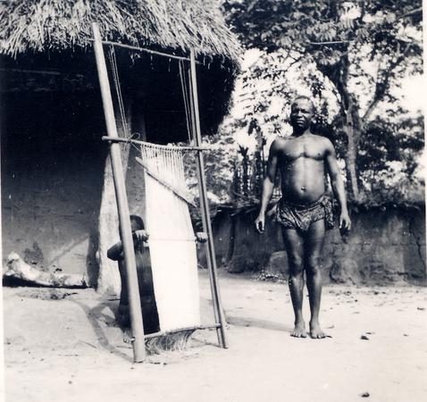 A person weaving on a vertical loom with a standing man in a loincloth.