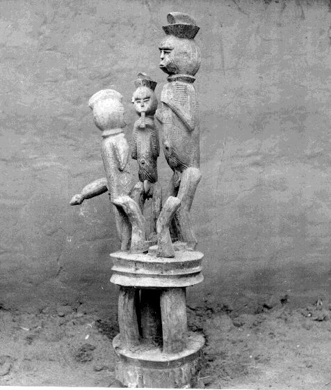 A multi-tiered vertical wooden sculpture with three carved figures on a pedestal.