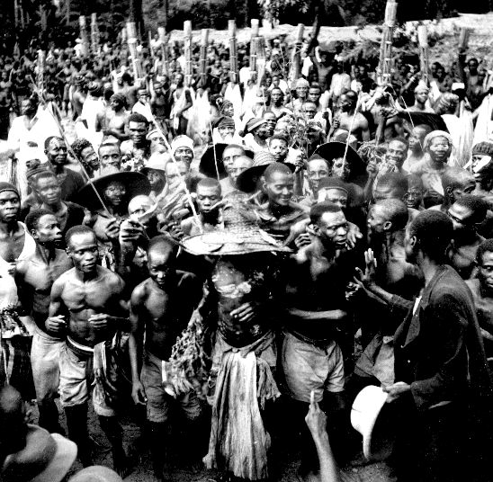 A photograph showing a group of individuals and a masked figure in raffia textiles during a boys' initiation in Nkporo.