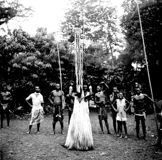 A figure in a fibrous mask and garment with a tall patterned pole during a boys' initiation in Nkporo.