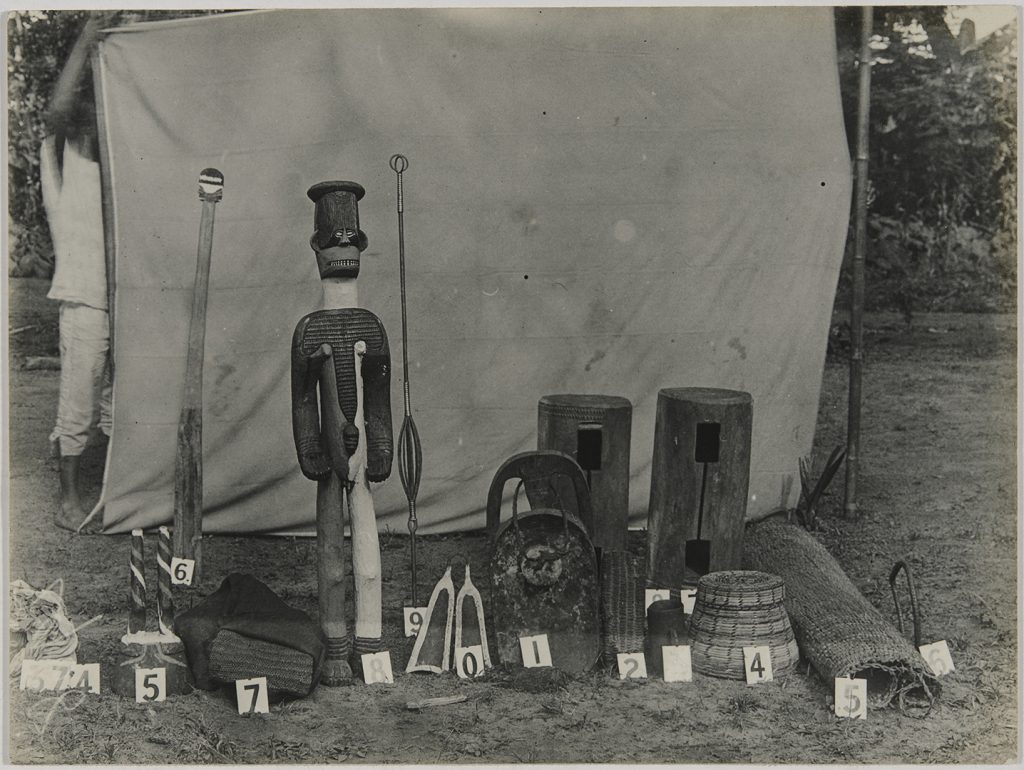 A monochromatic photograph of an arranged collection of Igbo artifacts including a tall Ngene figure and drums against a fabric backdrop.