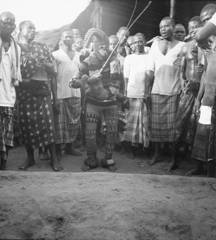 A black-and-white photograph of an Ekpe masquerader in Uzuakoli wearing a volumetric mask and fibrous costume.