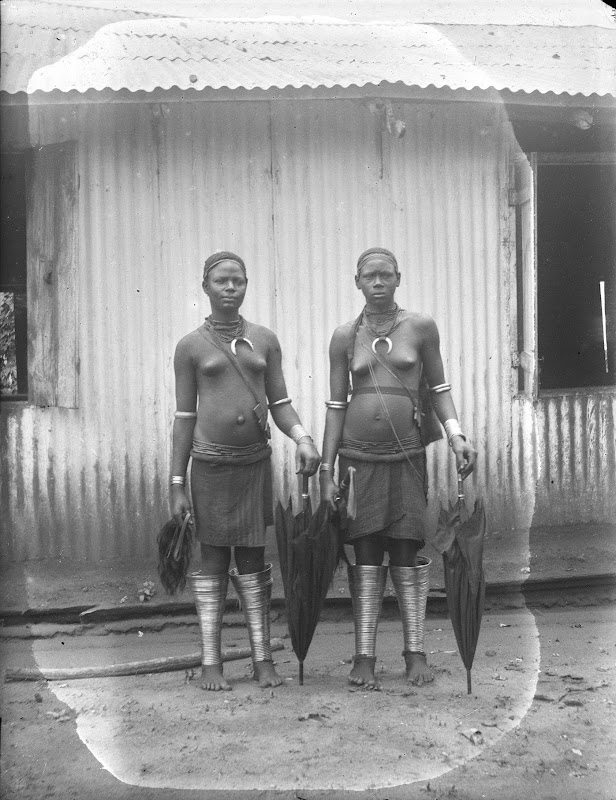 Two Igbo women wearing traditional necklaces and metallic leg guards.